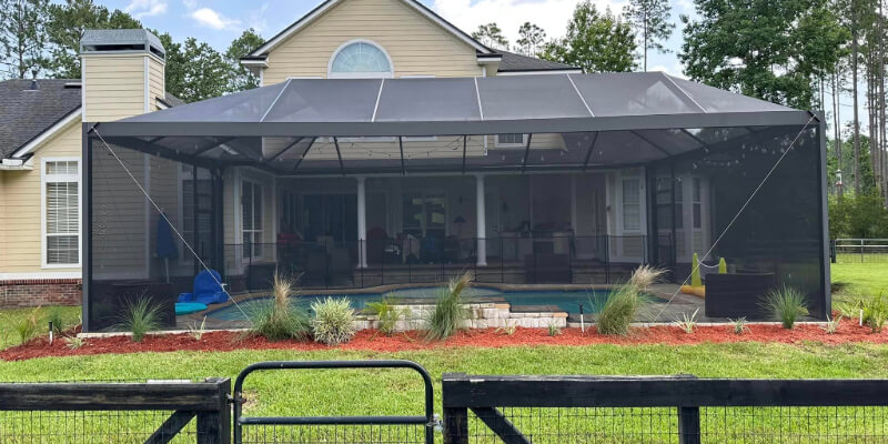 The picture shows a house with a big covered area for sitting and a pool. There are plants and red dirt around it. A black fence is in front. It looks like a nice place to relax outside.