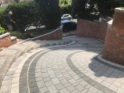 The driveway displays a professional circular paving pattern with precision-laid interlocking pavers in contrasting shades. Sharp edges and a clean integration with the surrounding brick walls highlight the meticulous craftsmanship.