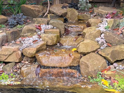 The image showcases a high-quality, professionally constructed water feature in a garden. Expertly arranged rocks create cascading waterfalls into a pond, surrounded by neat pebbles and vibrant plants, all indicative of skilled craftsmanship.