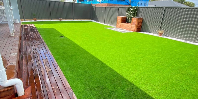 A vibrant green artificial turf backyard with a long wooden deck and corrugated metal fencing.
