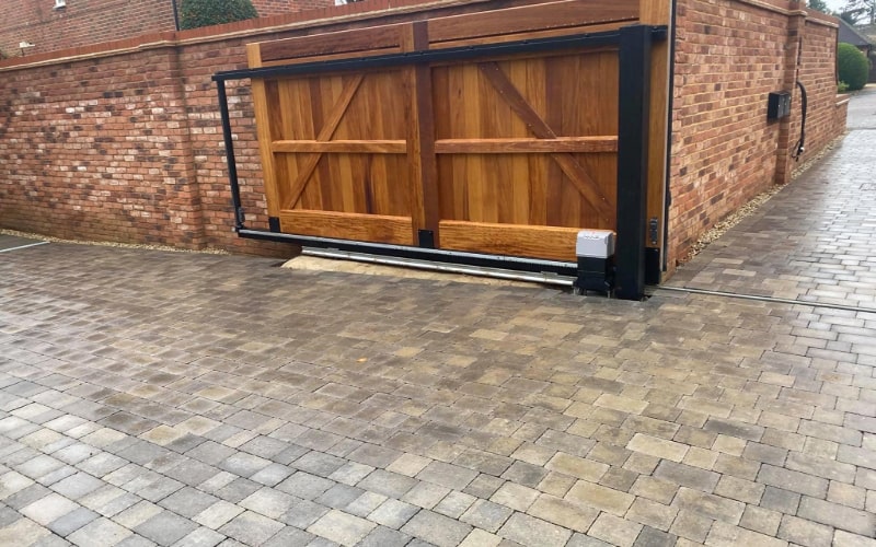 The image shows a driveway with professional paving composed of interlocking bricks in varying shades of brown. A large wooden gate and a brick wall are visible, creating a secure and private area.