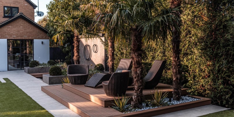 The image illustrates an elegant garden design with a wooden deck and wooden chairs, set among a backdrop of verdant palm trees and neatly trimmed hedges, exemplifying a modern, private outdoor space.
