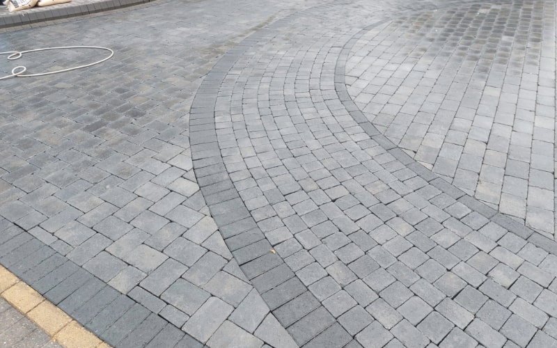 The image displays a professionally laid paving with dark grey bricks arranged in a sweeping curved pattern. The intricate brickwork shows attention to detail, suggesting a skilled paving job.