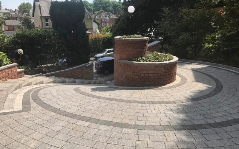 The image shows a driveway with professional paving. The paving stones are light grey and form a circular pattern around a brick planter. The area looks clean and well-maintained, with a car parked in the distance and trees around.