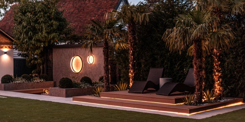 The image displays a modern garden design with a focus on minimalism and lighting. It features a manicured lawn, a raised deck with lounge chairs, and distinctive circular wall lights. LED strip lighting adds ambience, and strategically placed palm trees and shrubs create a private, tranquil outdoor space.