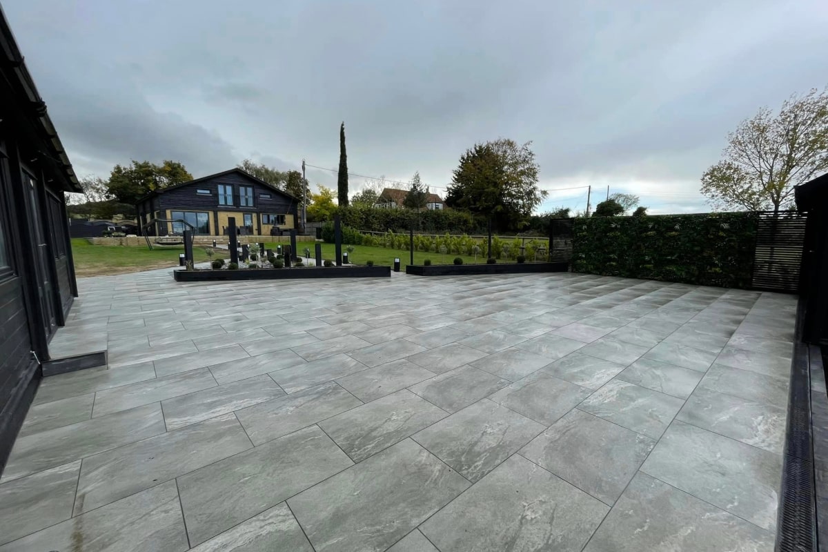 The image shows a large, open patio area with professional grey paving. The tiles are uniformly laid out and cover a spacious area, leading up to a modern house and lawn in the background.