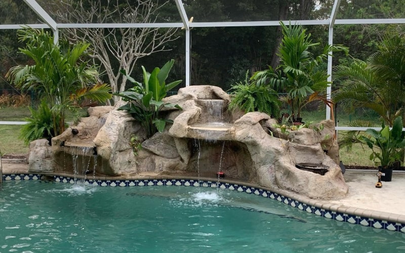 A professionally crafted rock water feature sits poolside, with water cascading gently from its tiered design into the pool below. The feature mimics natural stone formations and is surrounded by lush greenery, enhancing its authentic appearance.