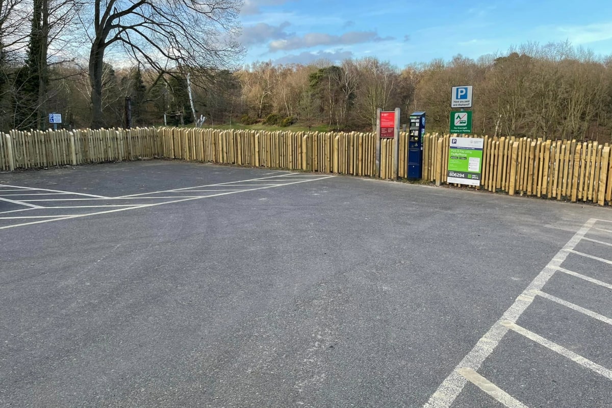 A well-maintained parking lot bordered by high-quality wooden fencing, showcasing precise, vertical slats evenly spaced and securely installed, indicating professional workmanship. The fencing stands uniformly along the lot's perimeter, complementing the neatly arranged parking spaces, with clear signage and a ticket dispenser adding to the organized setting.