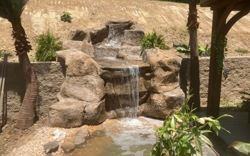 The image displays a professionally designed water feature that mimics a natural rocky waterfall. Expertly crafted, water smoothly cascades over multiple levels of stone, pooling into a serene pond at the base. Lush green plants and slender palm trees accent the feature, enhancing its tropical aesthetic. The backdrop of a bare soil hill contrasts with the water feature's verdant surroundings, emphasising its oasis-like appearance in the bright sunshine.