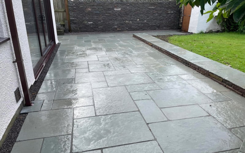 A neatly laid slate paving covers the patio area, featuring various sizes of rectangular and square tiles. The paving is expertly installed, providing a smooth and durable surface adjacent to a house and a well-maintained lawn.
